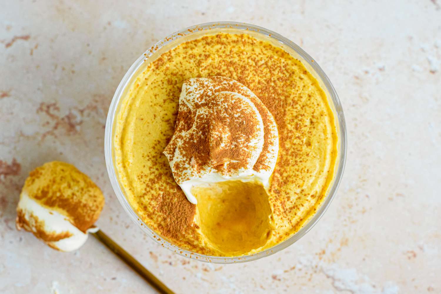 Overhead shot: 2-ingredient pumpkin mousse topped with whipped cream and a sprinkle of pumpkin spice, all in a sherbert glass. On the counter, a spoonful of mousse