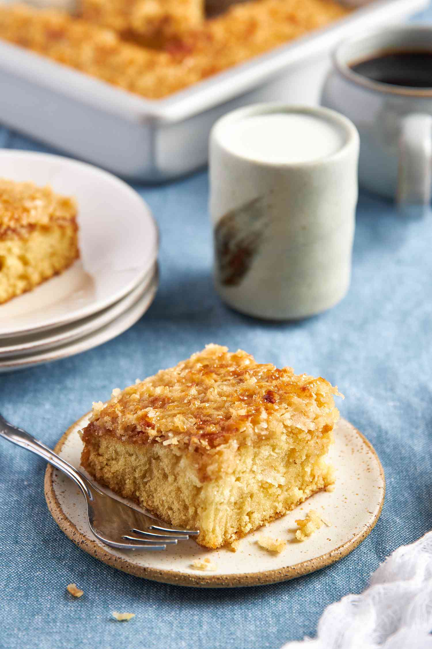 Slice of cake in a small plate with a fork, and in the surroundings, another slice on top of a stack of plates, a glass of cream, a mug of coffee, and a baking pan with more cake