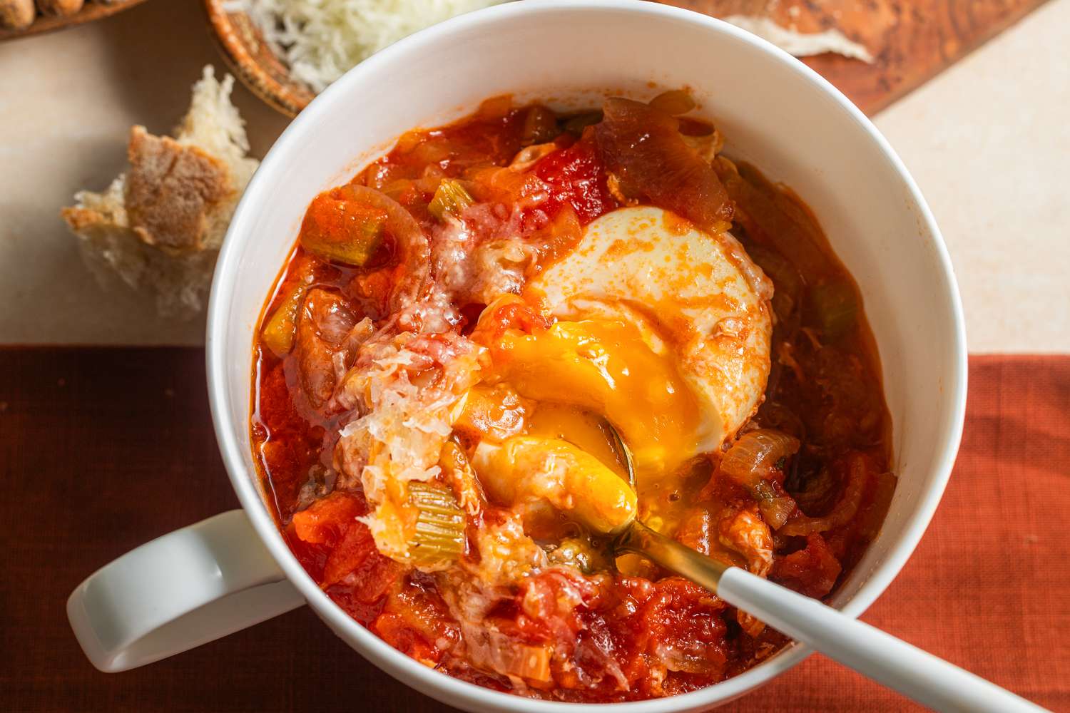 Overhead view of Acquacotta (Tuscan Soup) in a white bowl with a spoon plus bread on a cutting board and Pecorino romano cheese in a bowl to the side