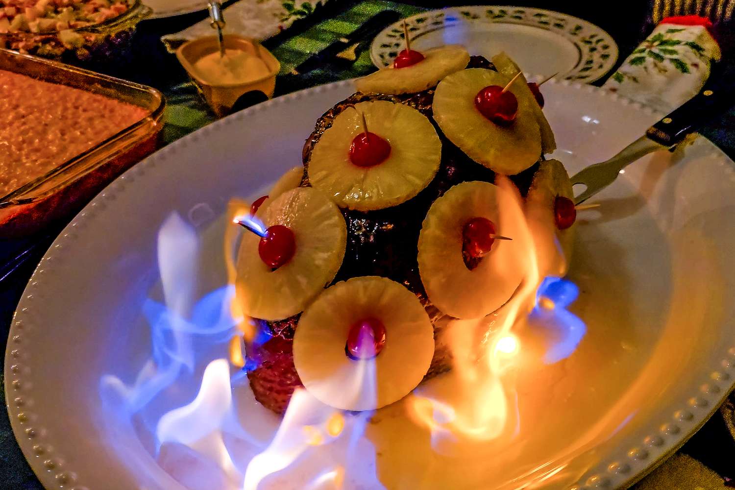 A cooked ham garnished with pineapple slices and cherries surrounded by flames on a serving plate