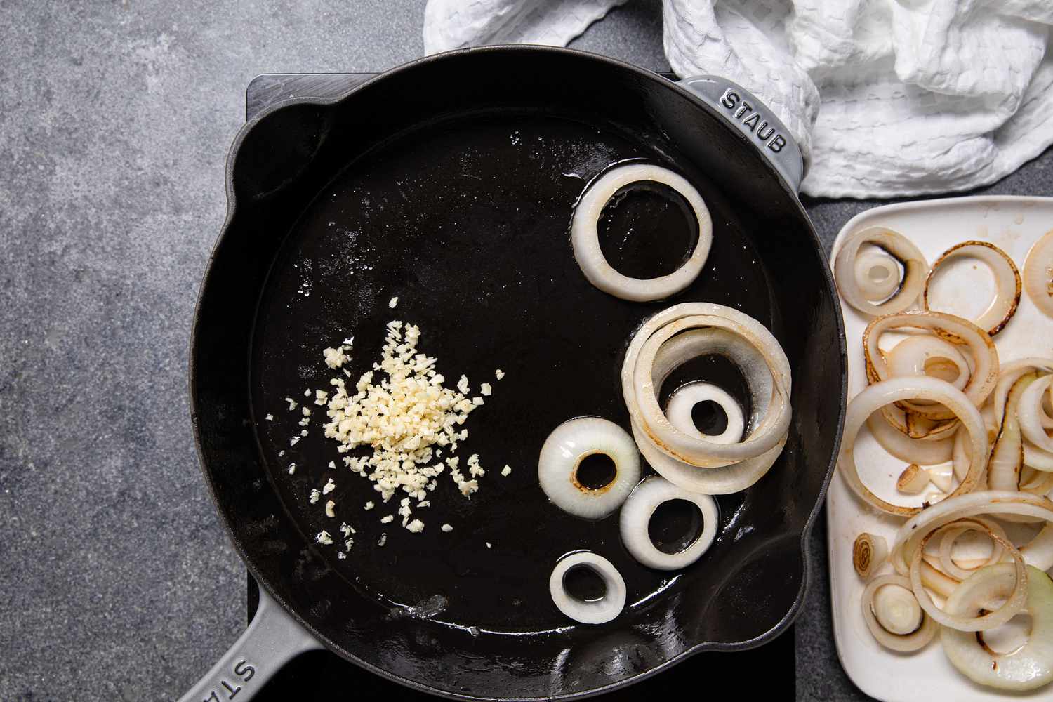 Minced Garlic and Some Onion Slices Cooking in the Cast Iron Skillet, and Next to It, a Plate of Cooked Onions and a White Kitchen Towel