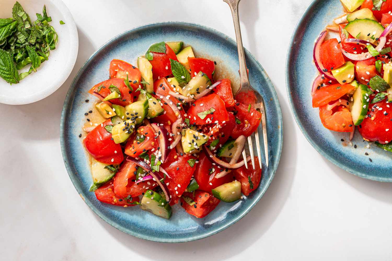 tomato salad topped with sesame seeds on two plates next to a small bowl of minced herbs