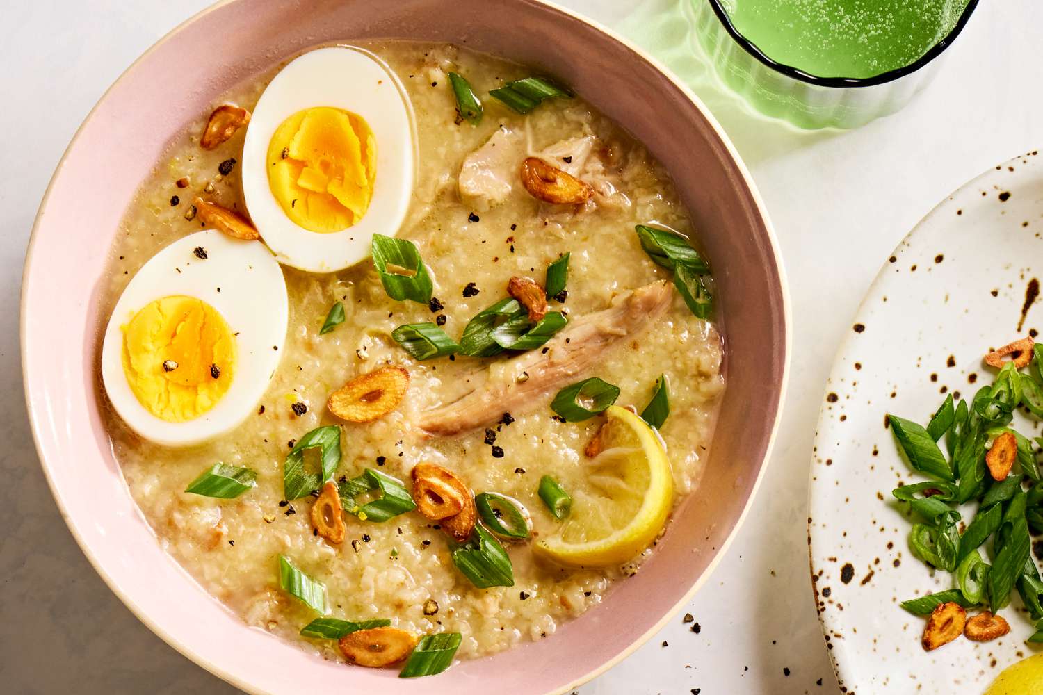 Overhead view of a pink bowl of Arroz Caldo with Chicken recipe next to a green glass and plate of lemon slices and scallions 