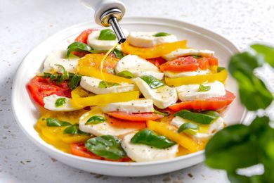 Olive Oil Drizzled Onto Platter of Caprese Salad With Tomatoes, Basil, and Mozzarella, and in the Foreground, a Basil Plant