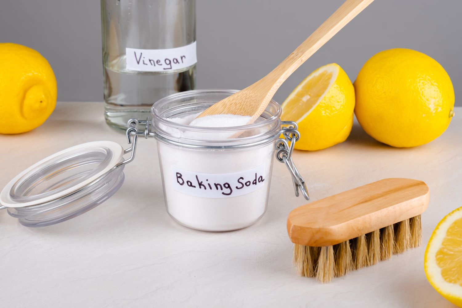 A glass jar of baking soda and a bottle of vinegar on a counter, with a cleaning brush and lemons scattered around