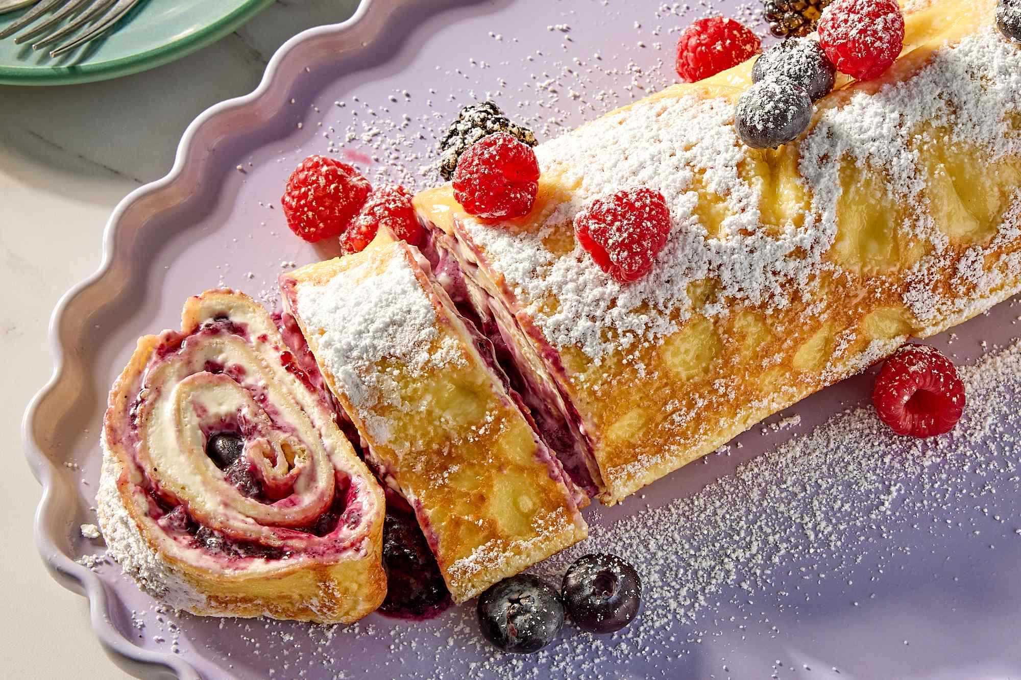 A rolled pastry filled with fruit and sprinkled with powdered sugar served with raspberries and blueberries on a lilac tray