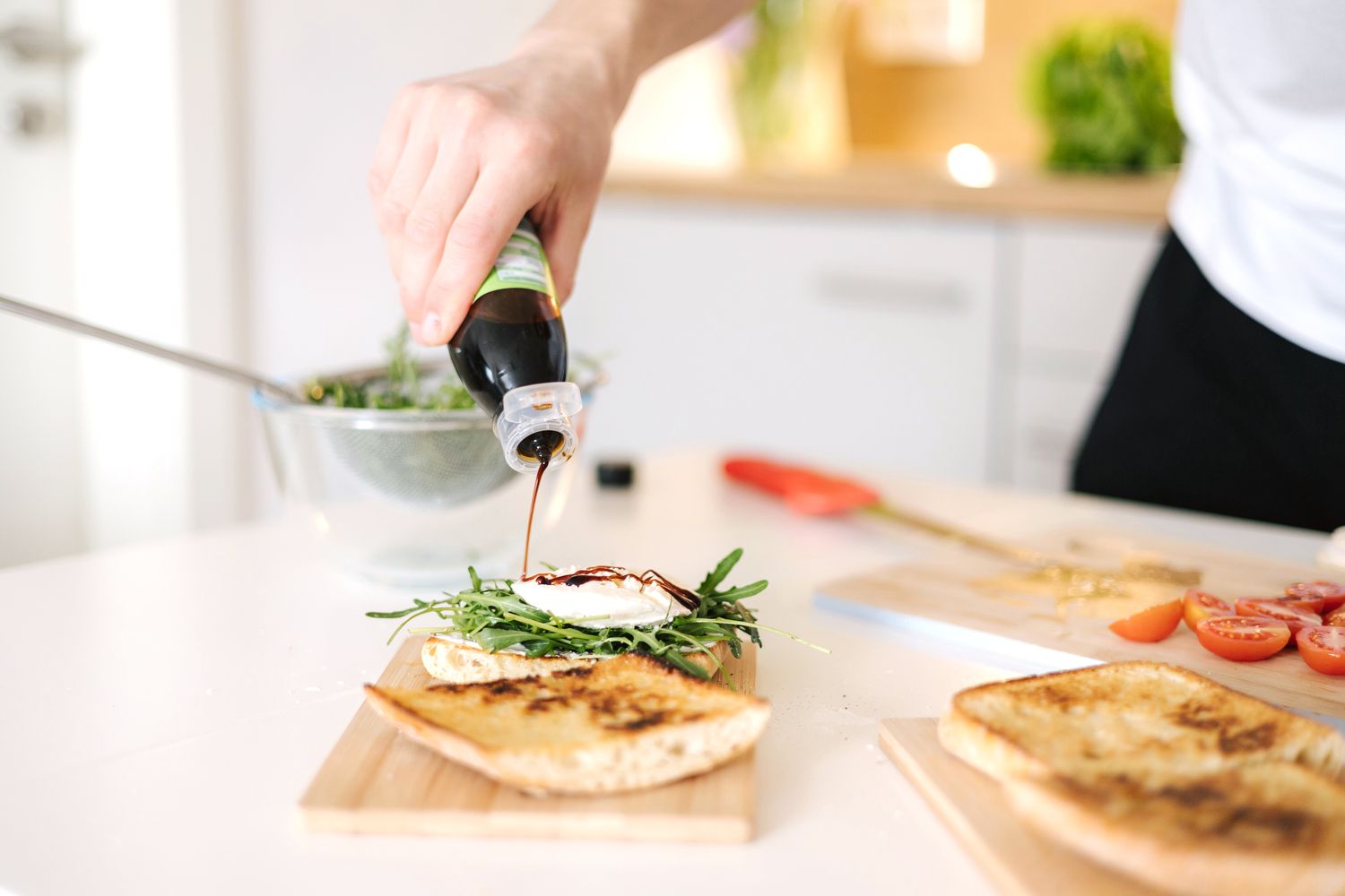 Person drizzling balsamic vinegar onto a bread slice topped with greens and a poached egg