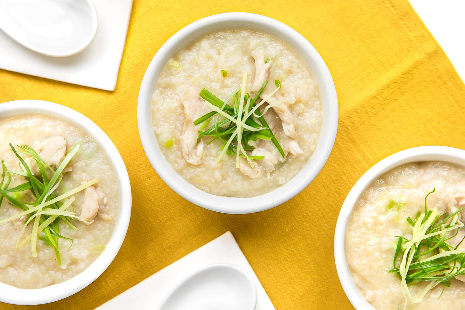 Bowls of Chicken Congee Topped with Sliced Ginger and Green Onions, Next to Soup Spoons, All on a Mustard Colored Kitchen Towel