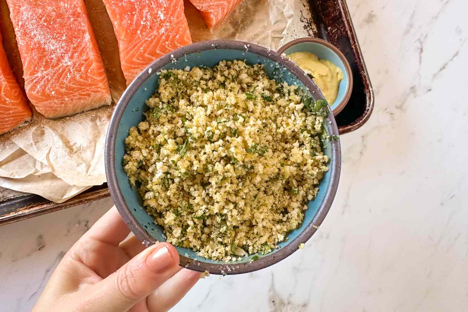 Overhead shot of a hand holding a bowl of seasoned breadcrumbs, with salmon fillets in the background