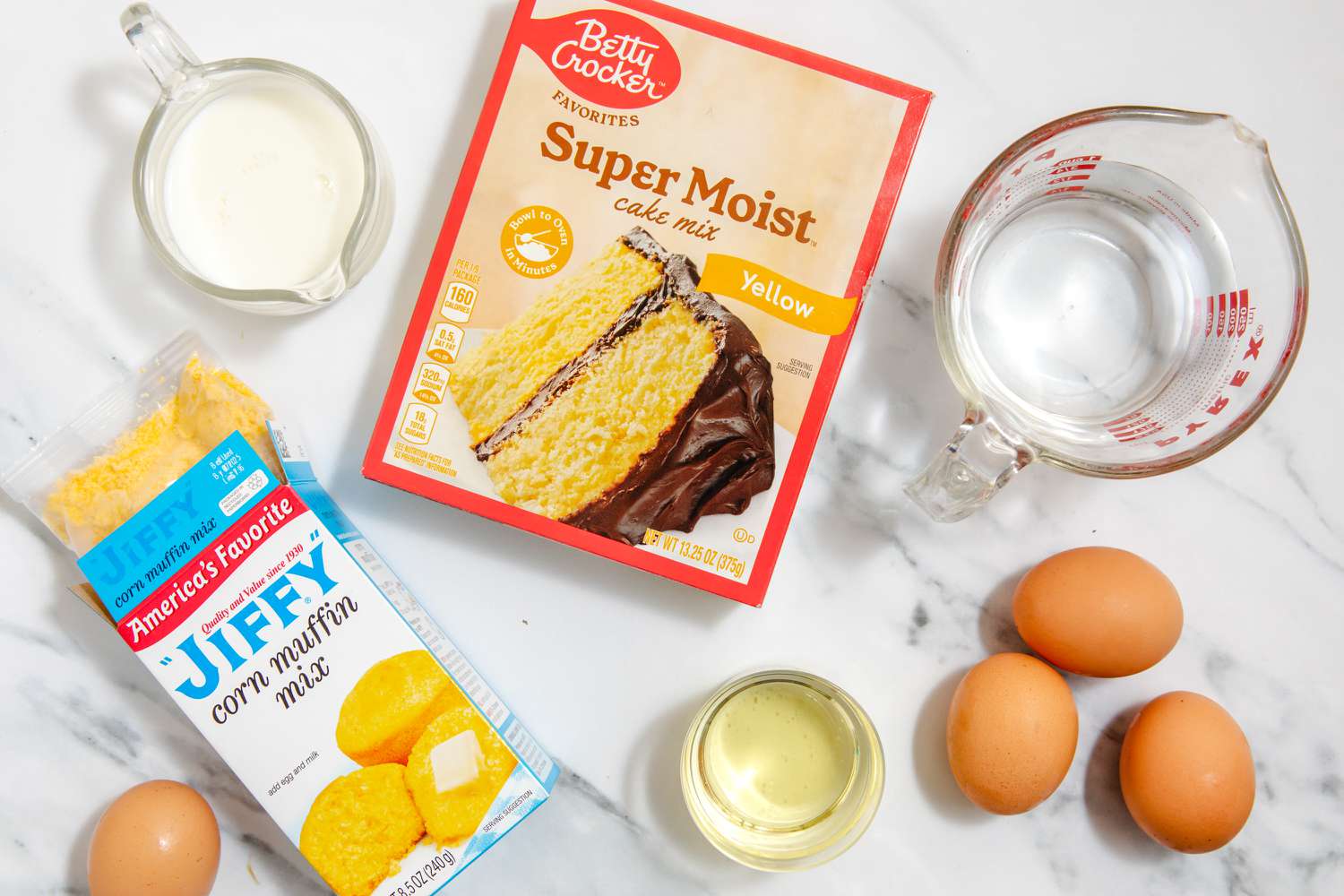 The ingredients for firehouse cornbread on a marble surface: yellow cake mix, Jiffy corn muffin mix, 4 eggs, oil, water, and milk