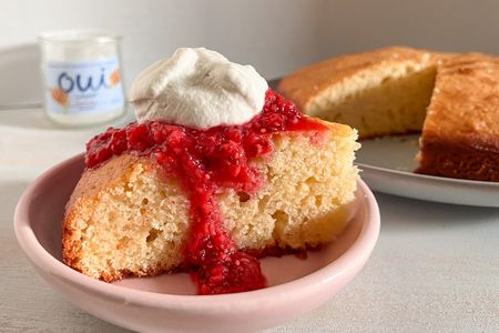 Slice of French yogurt cake with raspberry sauce and a dollop of whipped cream on a plate next to a cake pan with more and a bottle of Oui yogurt