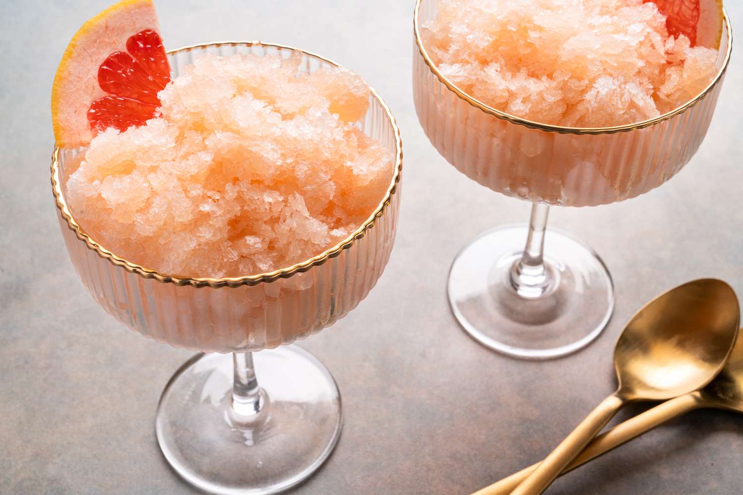 Two glasses of grapefruit granita garnished with grapefruit slices with spoons beside