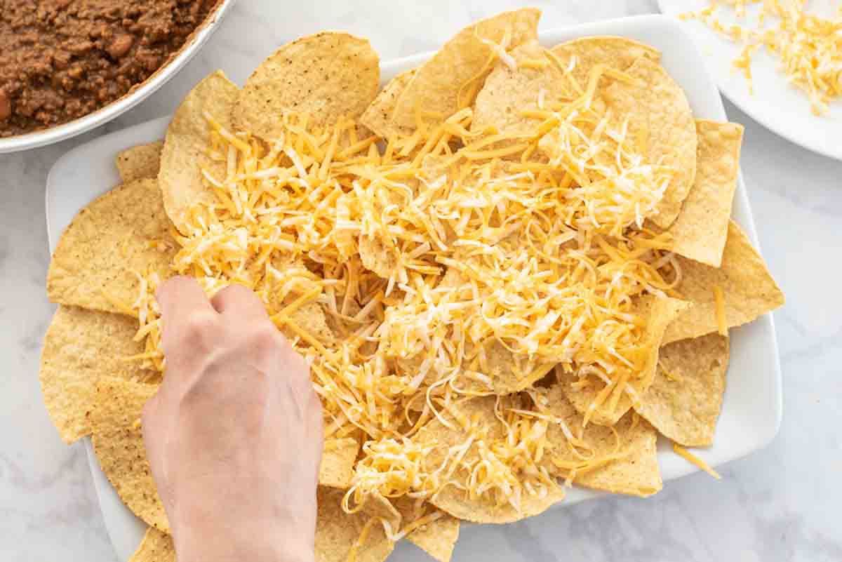 Tortilla chips on a white platter being sprinkled with shredded cheese with refried and ground beef mixture off to the side