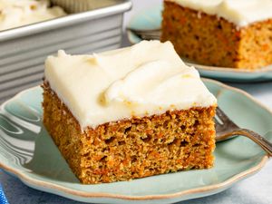 Carrot cake slice with cream cheese frosting on a plate, fork beside it, baking dish in the background.