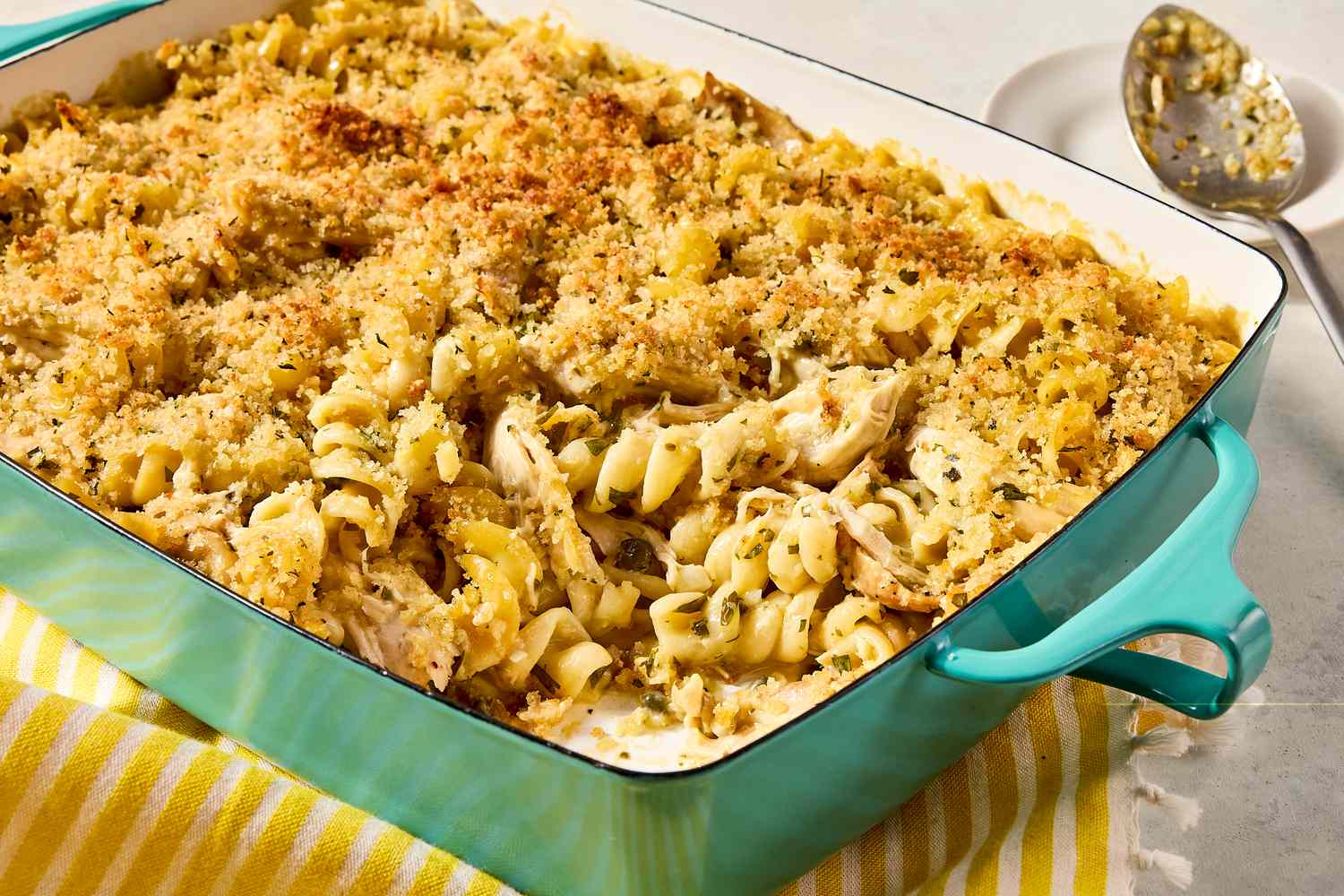 A casserole dish containing chicken piccata noodle casserole with a serving spoon nearby