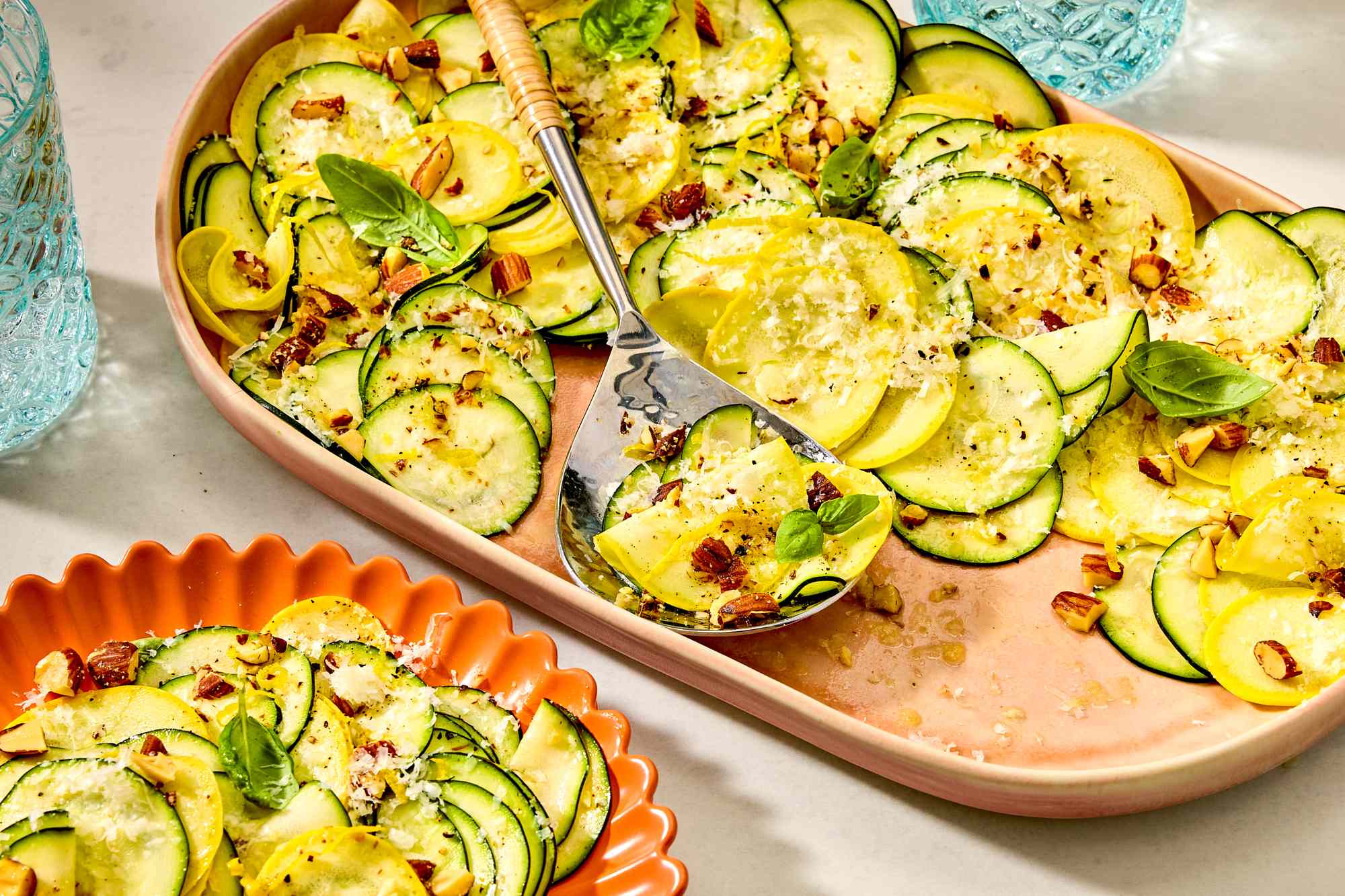 A platter and a plate with zucchini and squash salad sprinkled with nuts and cheese, garnished with basil leaves