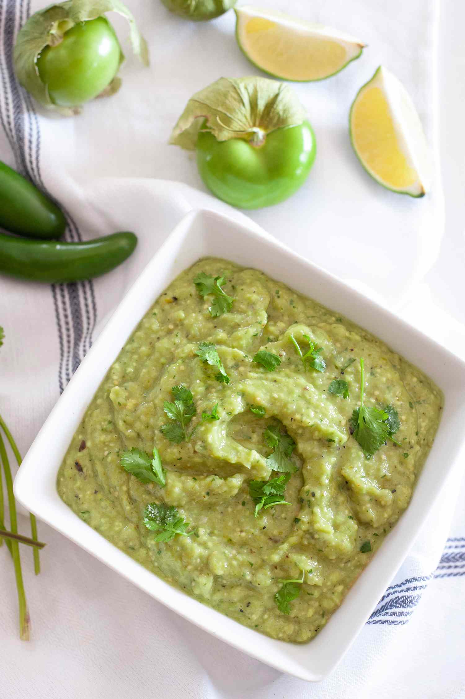 Bowl of Tomatillo Avocado Salsa Surrounded by Tomatillos, Lime Wedges, and Jalapeños