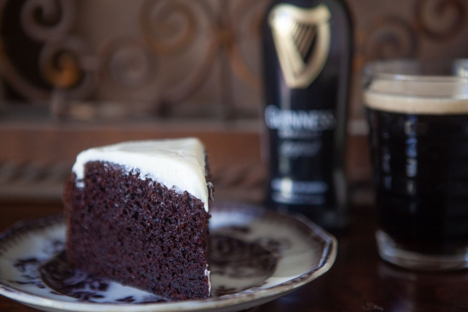 A Slice of Chocolate Guinness Cake on a Plate and in the Background, a Bottle of Guinness and More in a Glass 