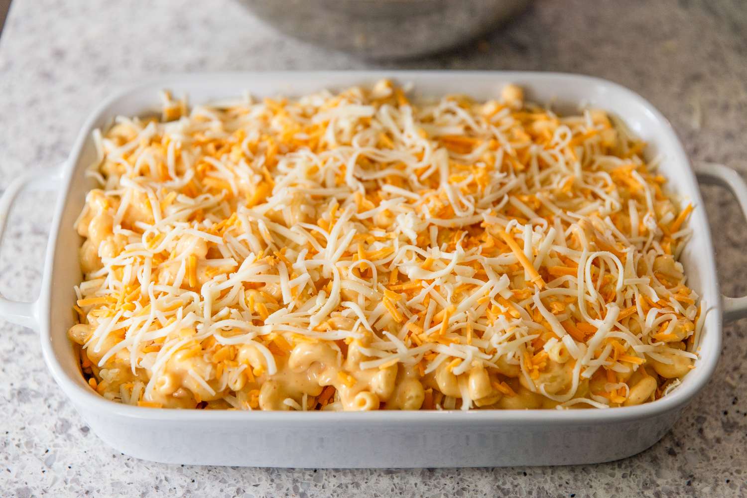 Angled view of a white baking dish of shredded cheese topping on the pasta and cheese sauce mixture from Baked Mac and Cheese recipe 