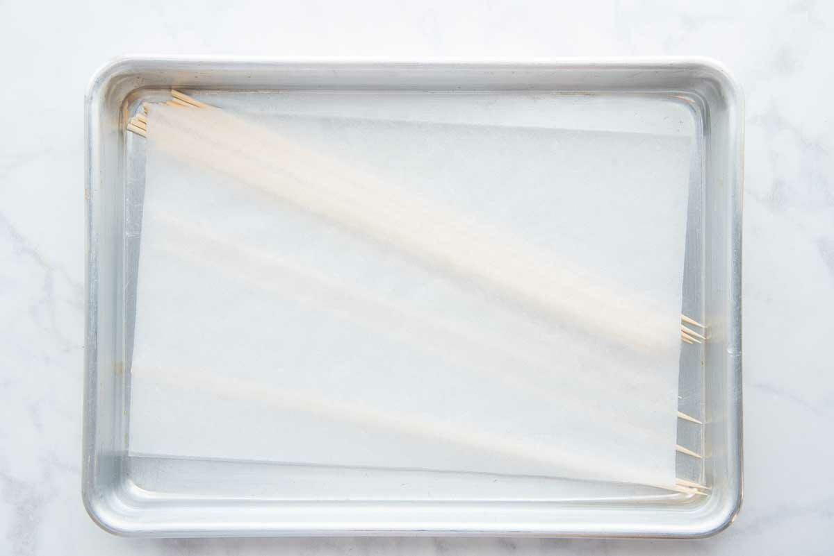 A quarter sheet pan has kebab sticks soaking in water with a piece of parchment set on top to keep the sticks submerged to make pork kebabs with grilled pineapple.