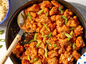 General Tsos chicken dish with sesame seeds and green garnish in a skillet served with rice on the side in a bluerimmed bowl