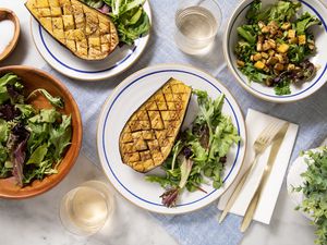 Table Setting (L to R): Small Bowl of Salt, Bowl of Salad Greens, Two Plates With Halved Eggplants and a Serving of Side Salad, Two Glasses, a Napkin With a Fork and Knife, a Bowl of Salad Greens Topped With Cubed Eggplant, and a Small Indoor Plant