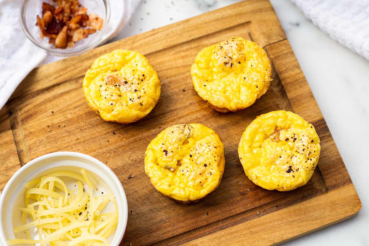 Bacon and Gruyère Egg Bites on a Wooden Board Surrounded by a a Small Bowl of Cheese and Another Small Bowl with Bacon Bits