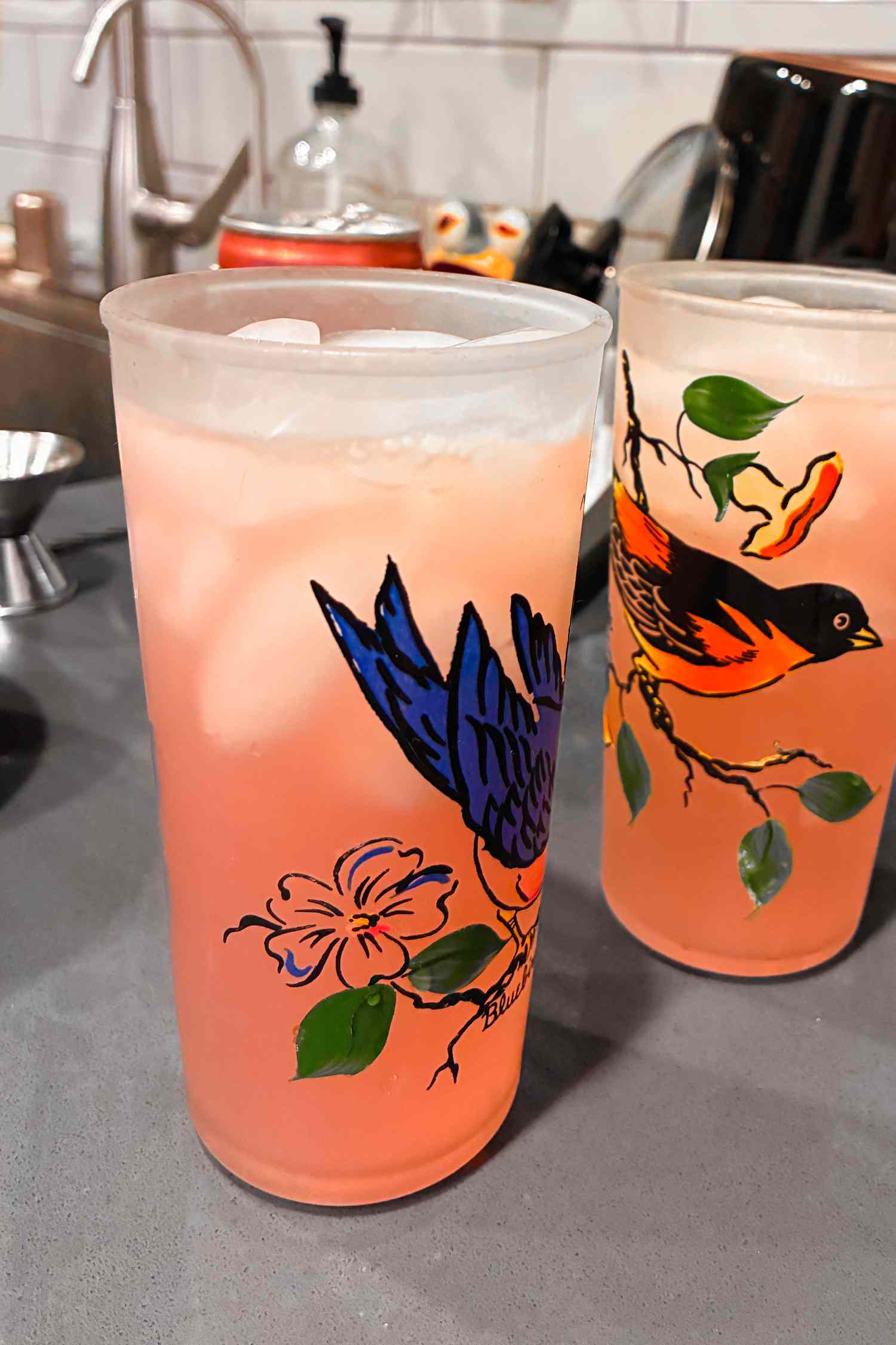 Two paloma cocktails in glasses with birds painted on them sitting on a kitchen counter
