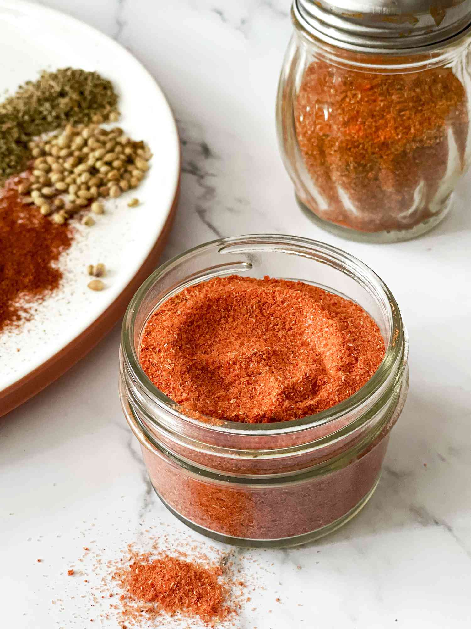 Jar of Homemade Sazon Spice Mix with Shaker and Plate of Spices in Background