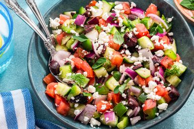 Bowl of Easy Greek Salad With Utensils, and Next to It, a Glass of Water, a Small Bowl of Herbs, and a Kitchen Towel