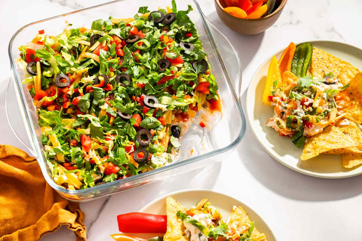 Layered nacho dip in a glass dish surrounded by tortilla chips and sliced vegetables on plates