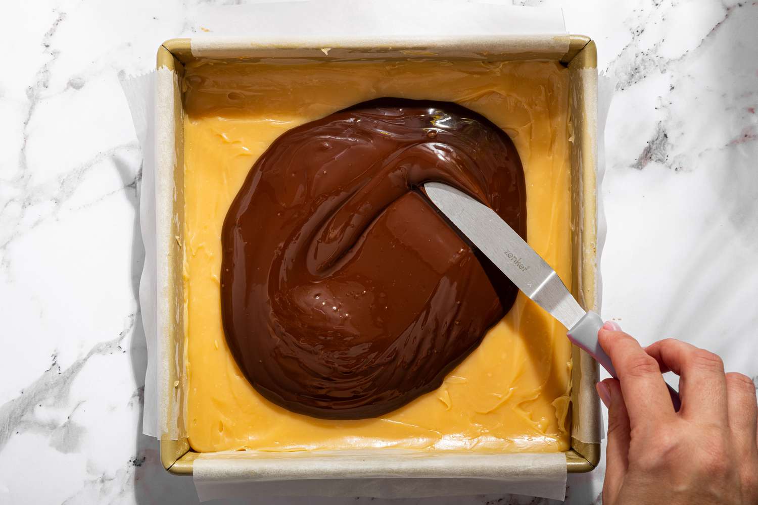 Overhead view of a baking pan showing the spreading of the melted chocolate onto the caramel layer for Millionaire Shortbread recipe