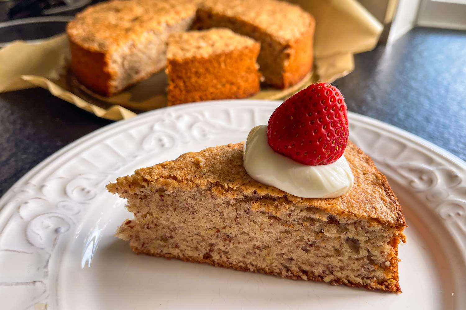 Slice of almond cake topped with whipped cream and a strawberry on a white plate, whole cake in the background