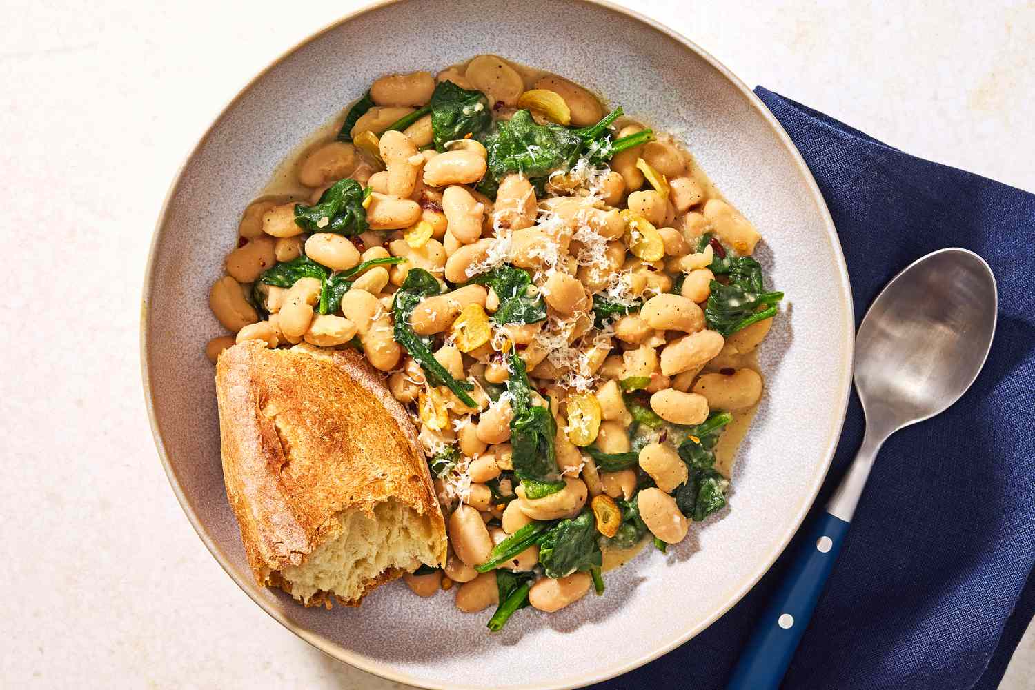 Overhead view of a bowl of beans and greens with a chunk of bread all next to a spoon and resting on a blue cloth napkin