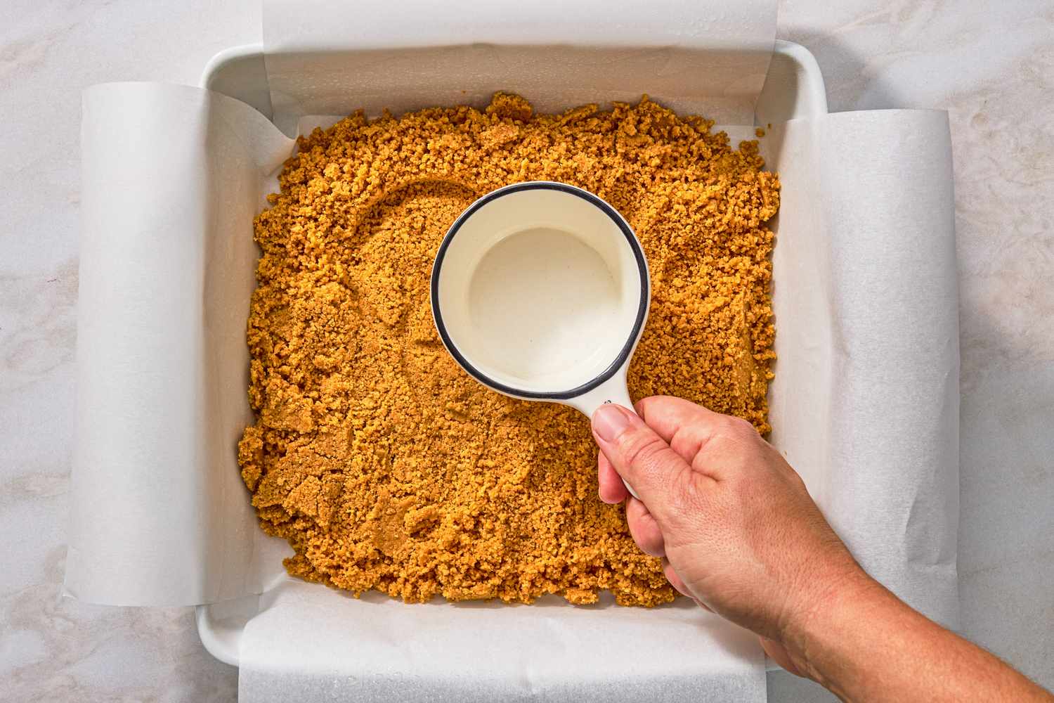 hand using cup to press cookie crumbs into parchment lined baking dish for Chocolate-Peanut Butter Delight