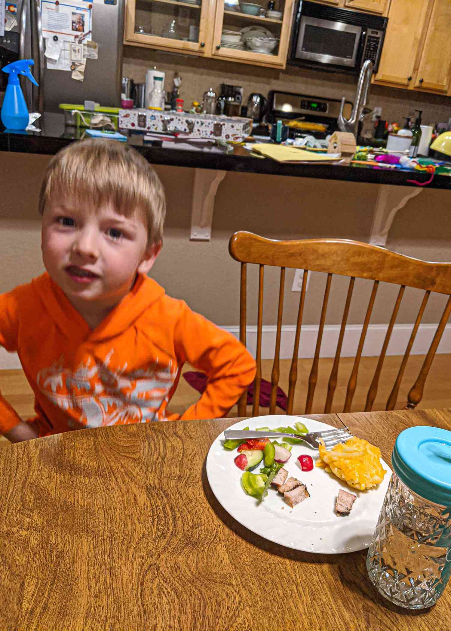 Preschool boy sitting at the kitchen table with a white plate of food in front of him The plate has a scoop of cheesy funeral potatoes, chopped ham, a salad and a fork. The kitchen is in the background of the picture.
