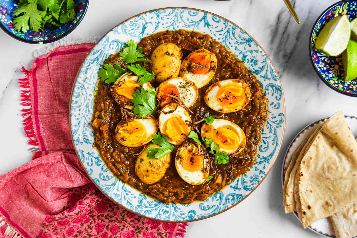 Plate of Egg Masala Curry on a Kitchen Linen, Surrounded by a Plate with Roti, a Bowl with Lime Wedges, and a Bowl with Cilantro