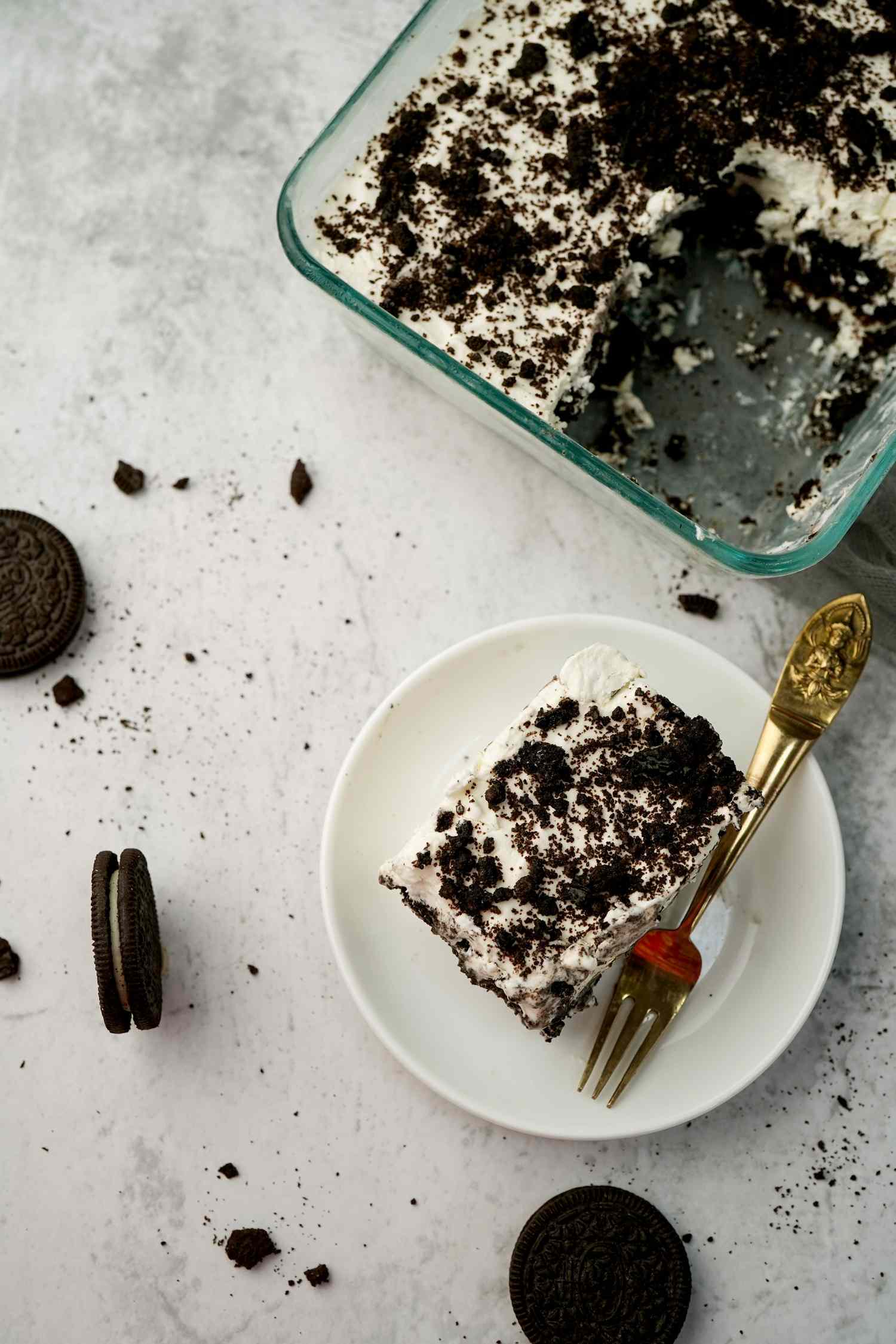 A serving and a tray of cookies and cream icebox cake with a fork on a white surface