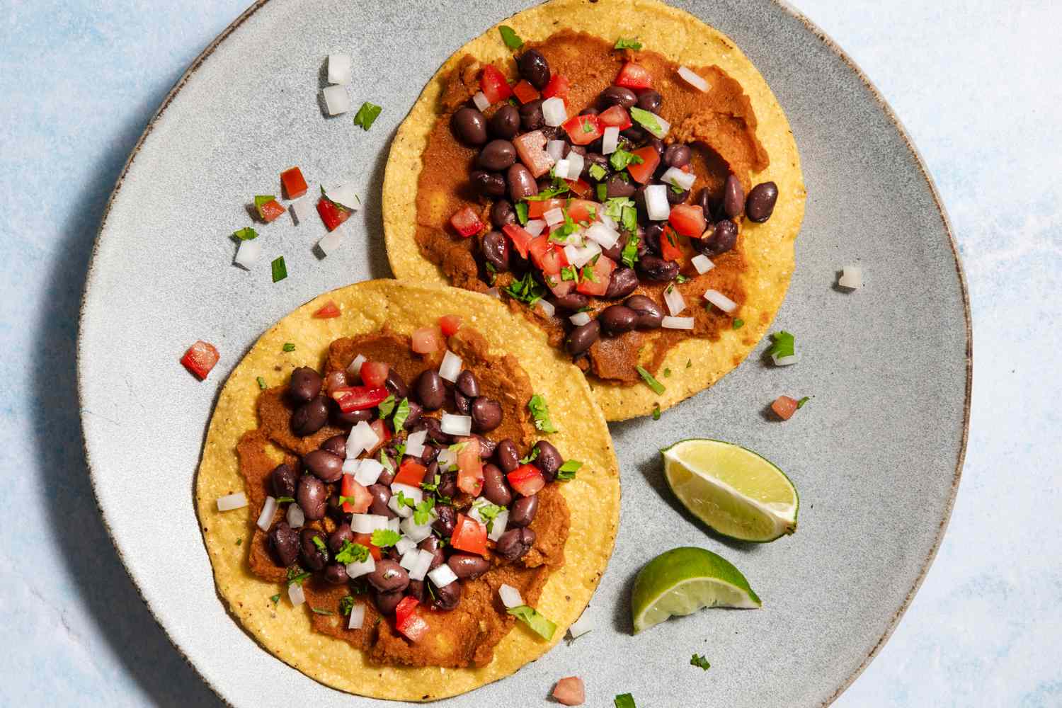 Two tostadas topped with black beans diced vegetables and herbs served with lime wedges on a plate