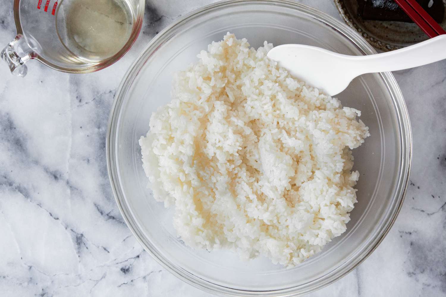 Sushi Rice in Large Bowl with a Rice Paddle Next to Measuring Cup with Vinegar Mixture and Bowl with Discarded Kombu