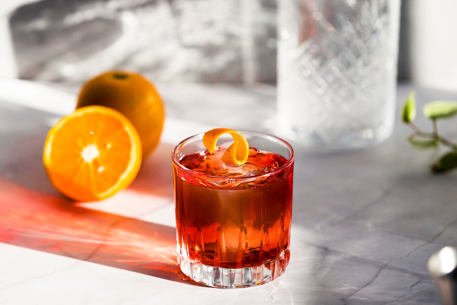 A negroni cocktail garnished with an orange peel strip, with oranges and glassware in the background