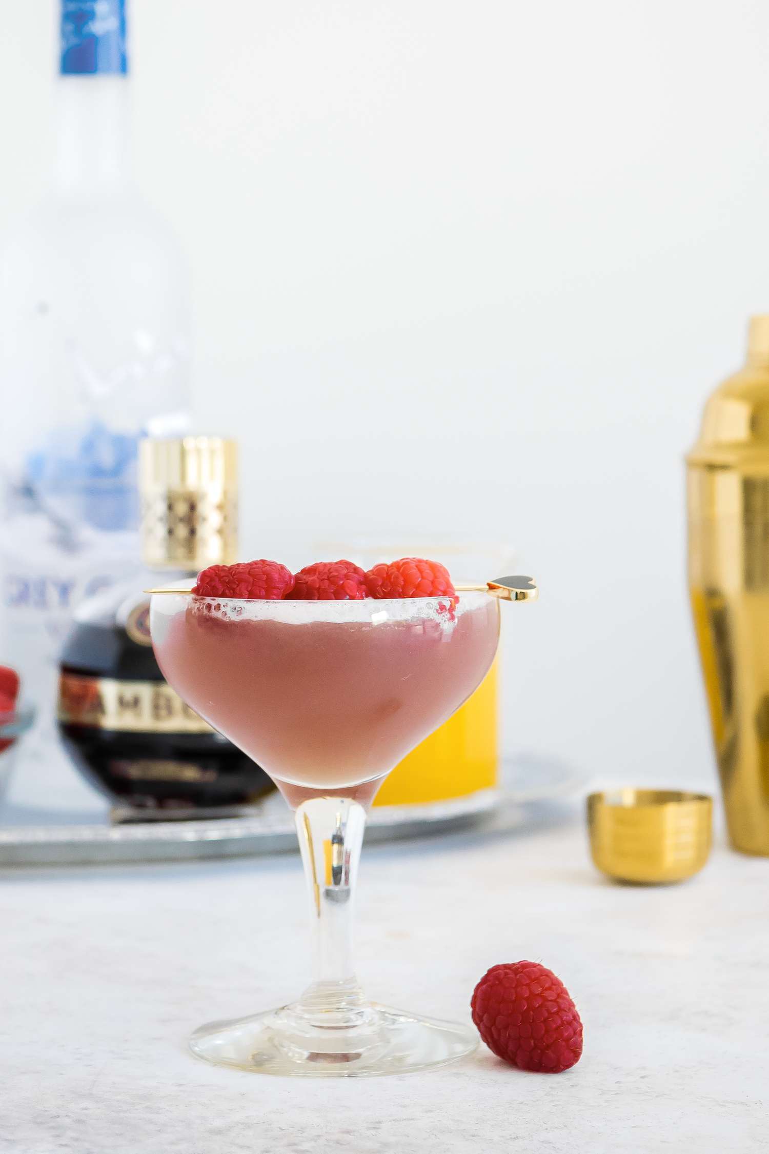 French Martini in a Glass Garnished with Raspberries and Surrounded by Shaker and a Tray with a Bowl of Raspberries, Vodka, Chambord, and Pineapple Juice 