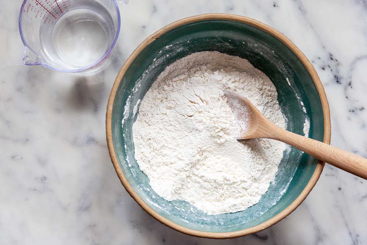 A wooden spoon is inside a bowl with flour inside. A measuring cup of water is to the left of the bowl.