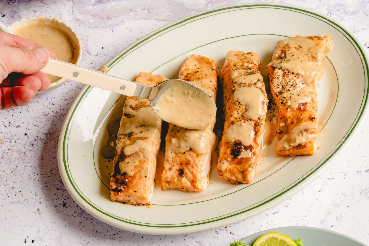 Four pieces of salmon being drizzled with a creamy Caesar dressing with a spoon on a serving plate