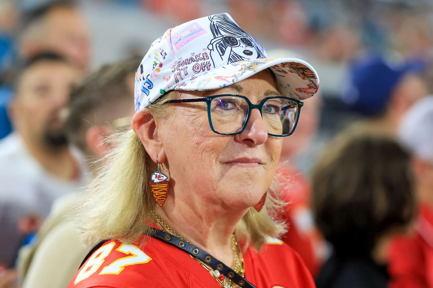 A woman wearing a decorated sports cap and Chiefsthemed earrings in a crowded setting with her attention seeming directed towards an event