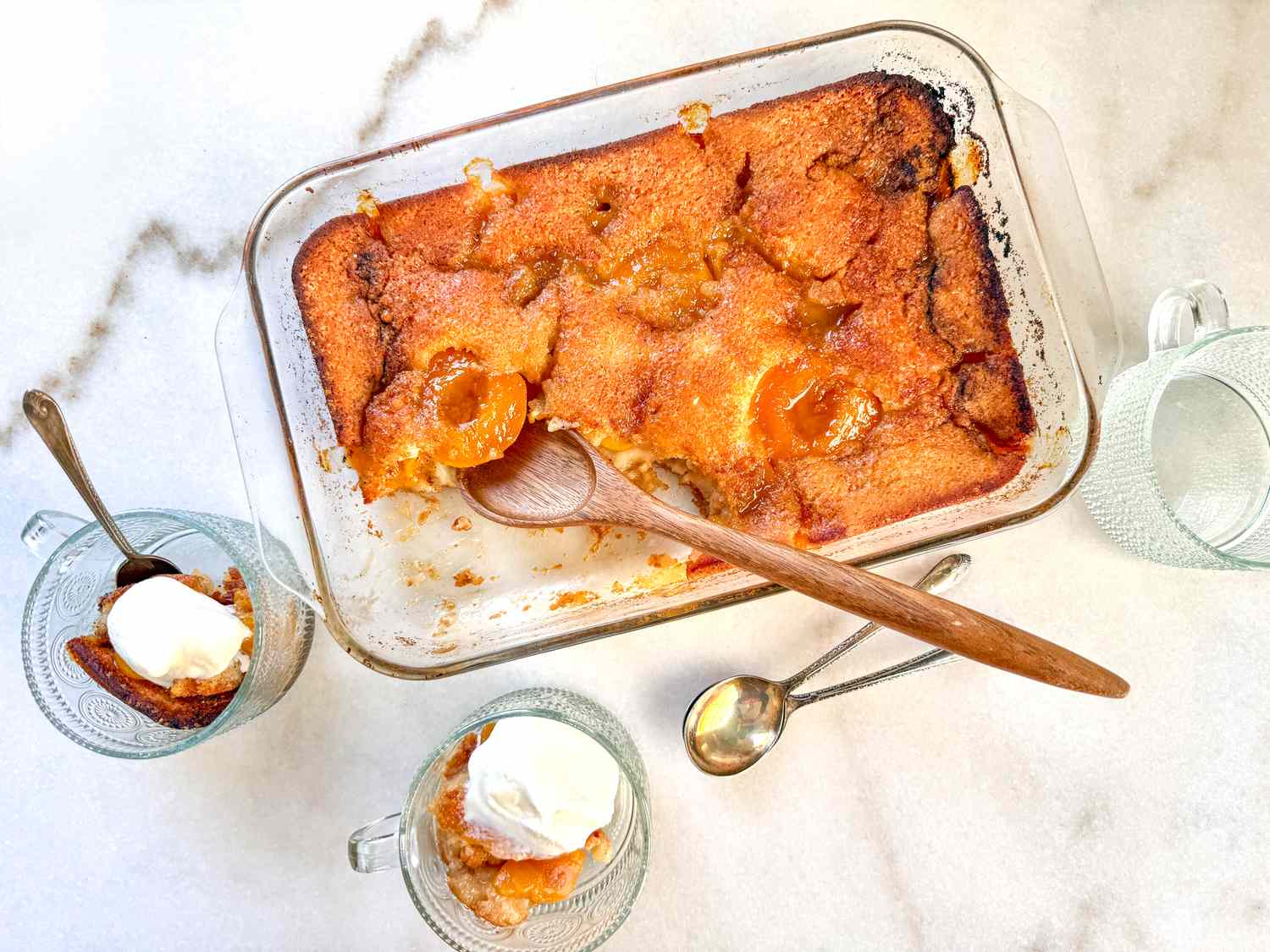 Peach cobbler in a baking dish, served in small bowls with spoonfuls of cream