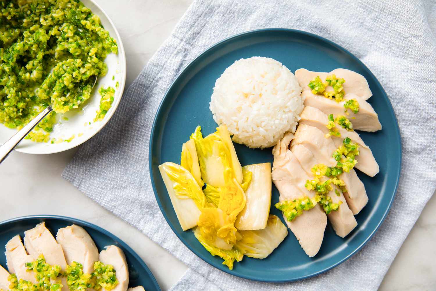 A blue plate with sliced chicken topped with scallion sauce, a portion of rice, and lettuce. A bowl of scallion sauce sits nearby