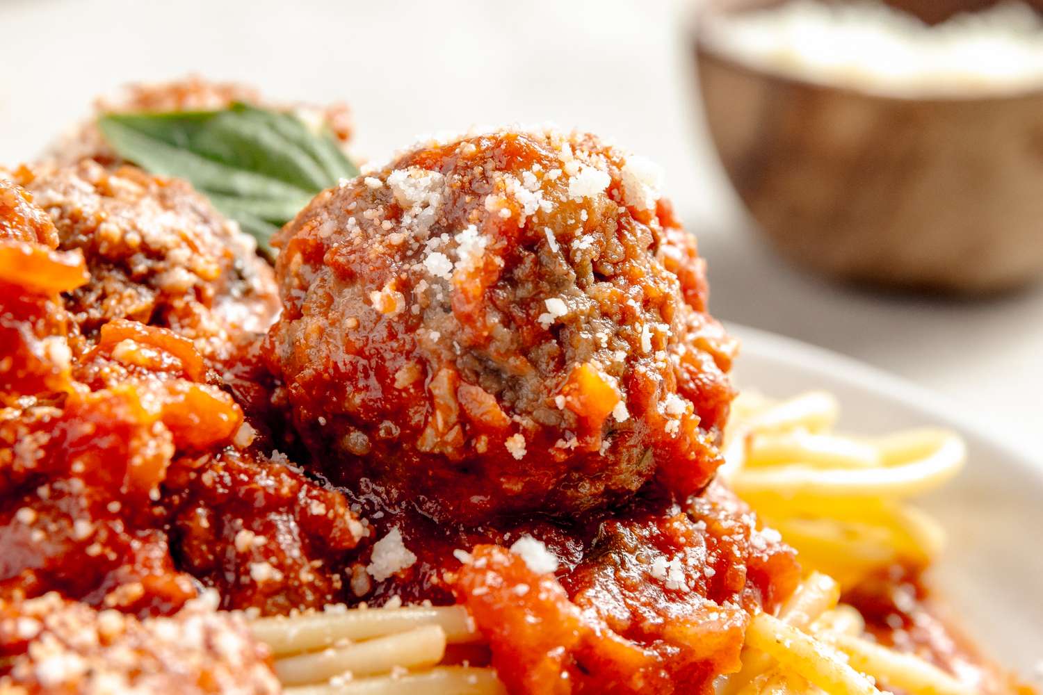 Closeup of a meatball on top of spaghetti with marinara sauce garnished with grated cheese and basil leaves