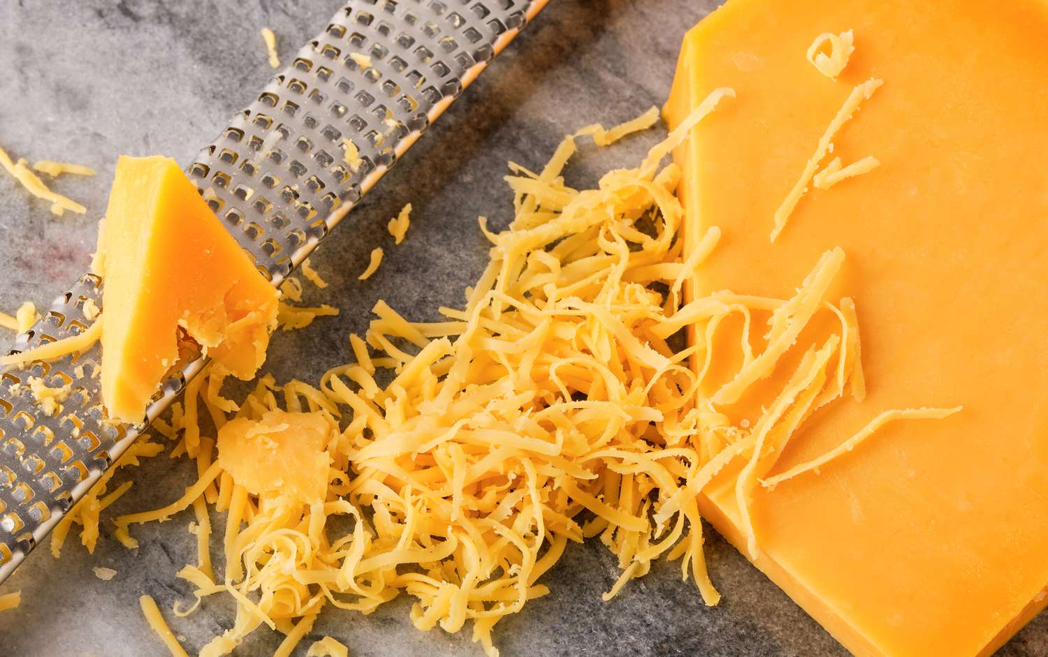 Overhead view of a large block of cheddar cheese, a shredder and pile of shredded cheese on a marble countertop