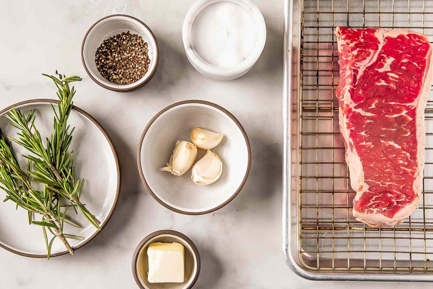 Ingredients for cooking steak including raw meat, rosemary, garlic, salt, butter, and pepper